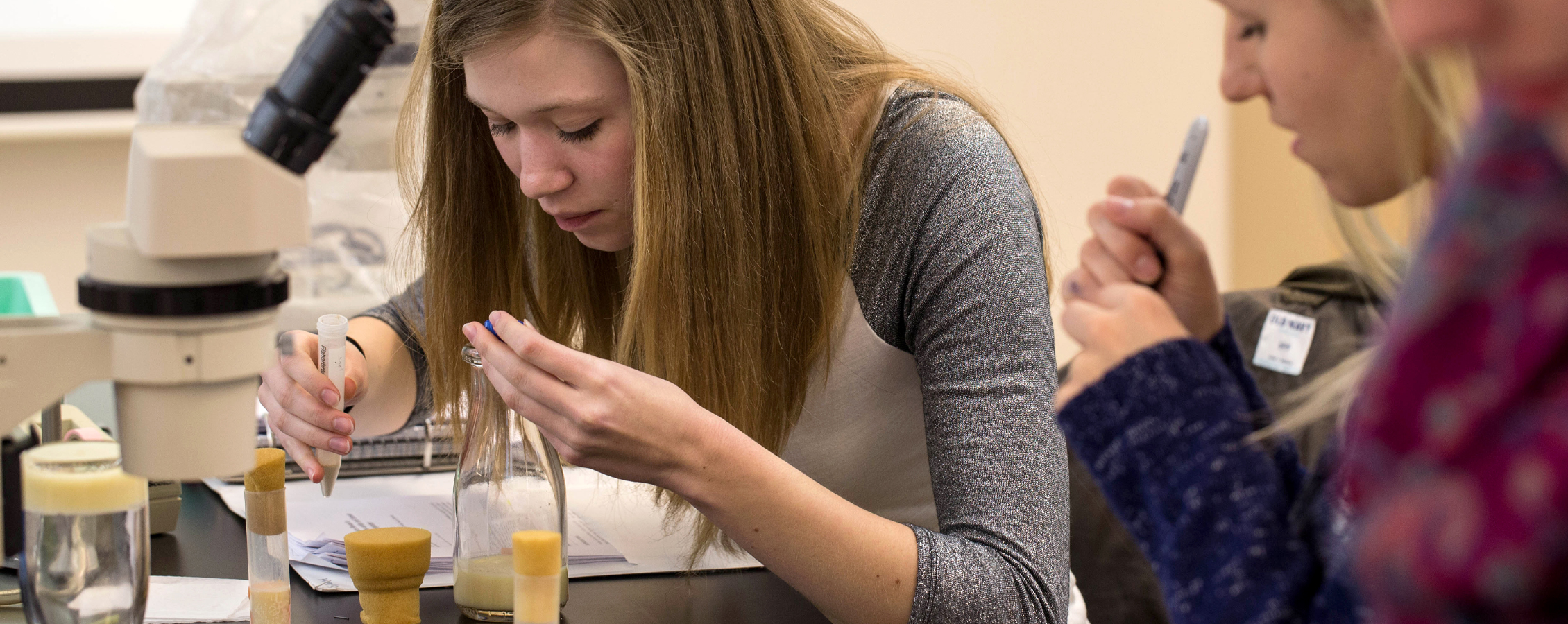 A student inspects something in a vial next to a microscope.