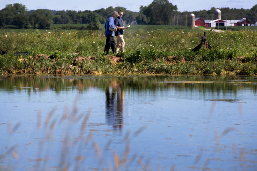 Two people walk by a waterway with a farm in the background.