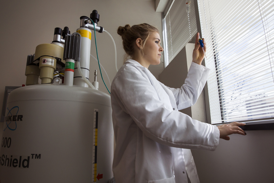 A student in a lab coat looks at a vile by the window.