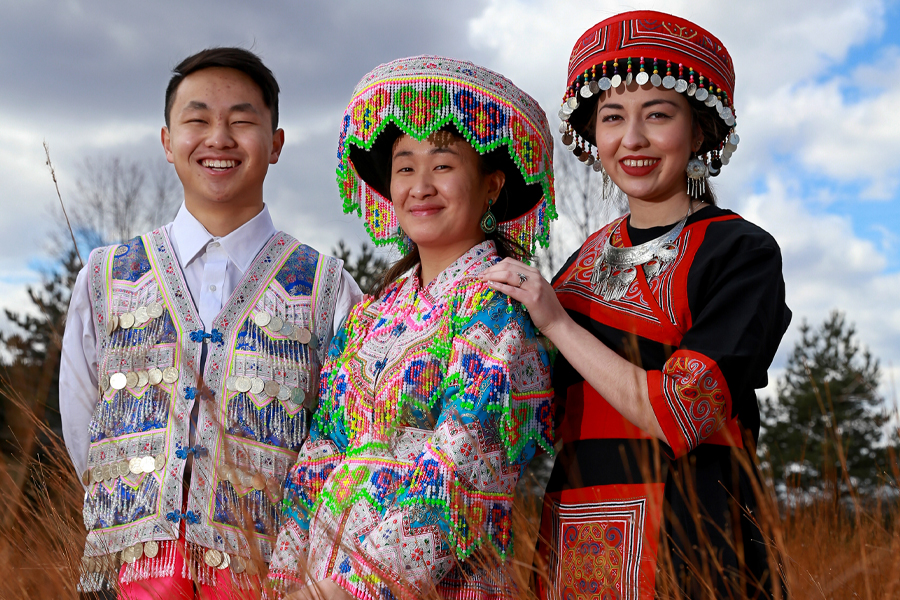 Three Asian students model traditional Hmong clothing, jewelry and story cloth.