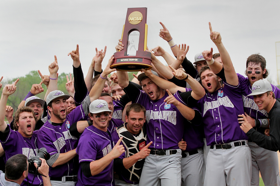 Men's baseball cheers and holds up an NCAA trophy on a baseball diamond.