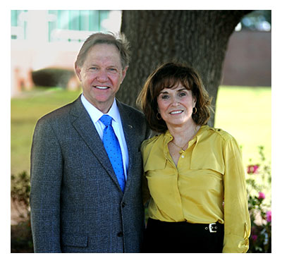 Two people stand outside by a tree and smile at the camera.