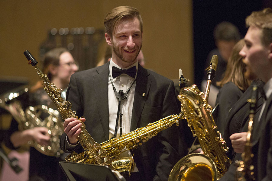 A student wearing a suit and tie smiles while holding a saxophone.