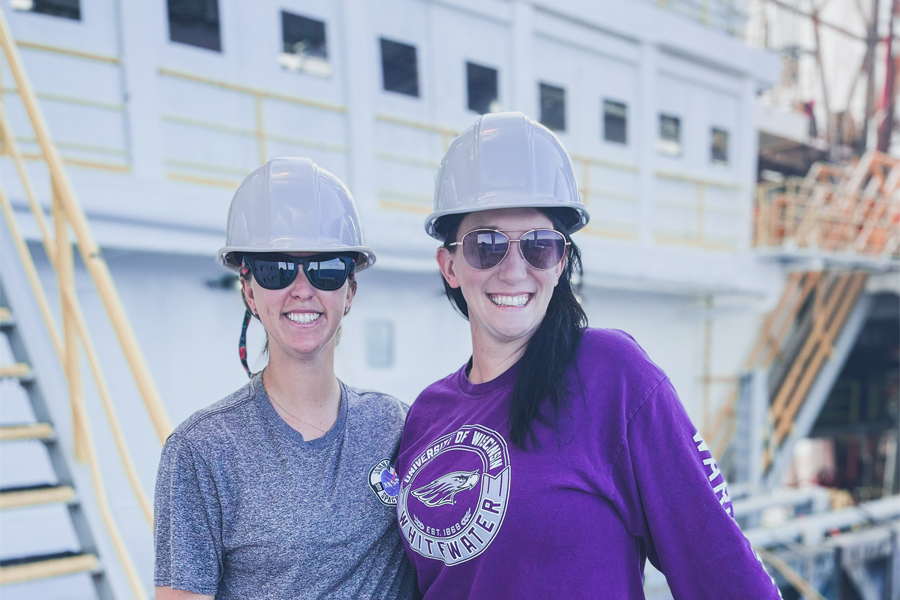 Two young people wearing hardhats, smile at the camera with a large ship in the background.