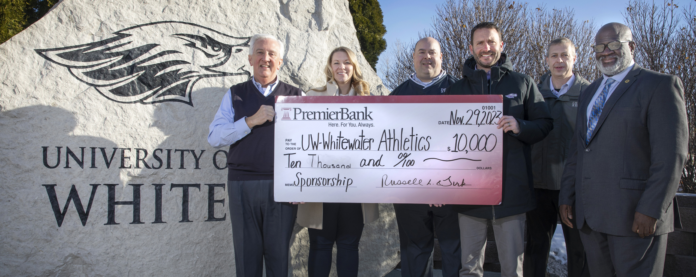 Six people hold a large check and stand by a UW-Whitewater Warhawk monument.