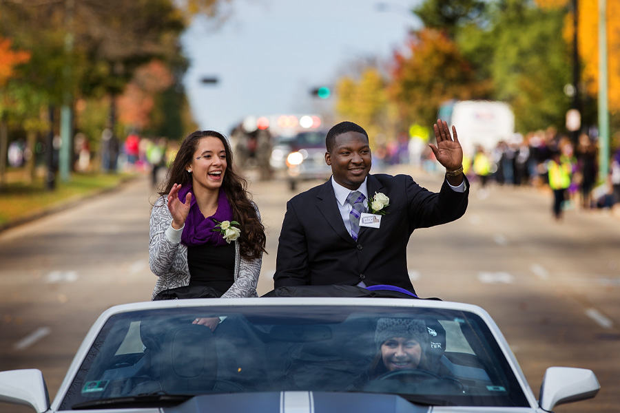 Two people wave from a convertible car during a Homecoming parade.