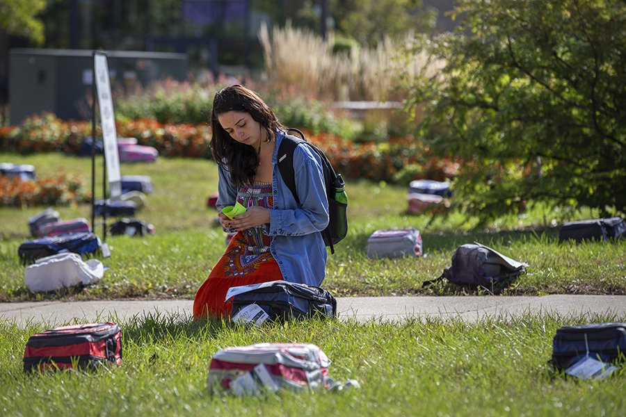 A person kneels on the grass reading a note on a backpack.