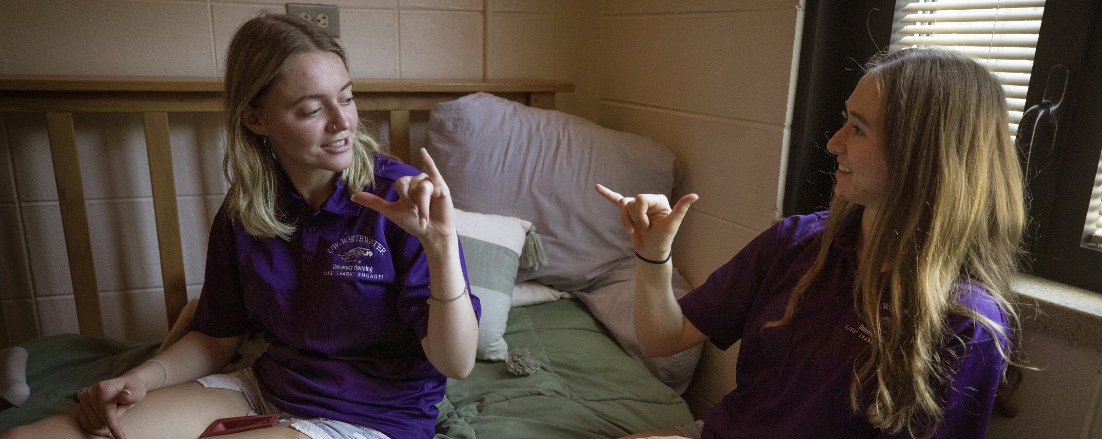 Two people in purple shirts sit on a bed together and sign with their hands.