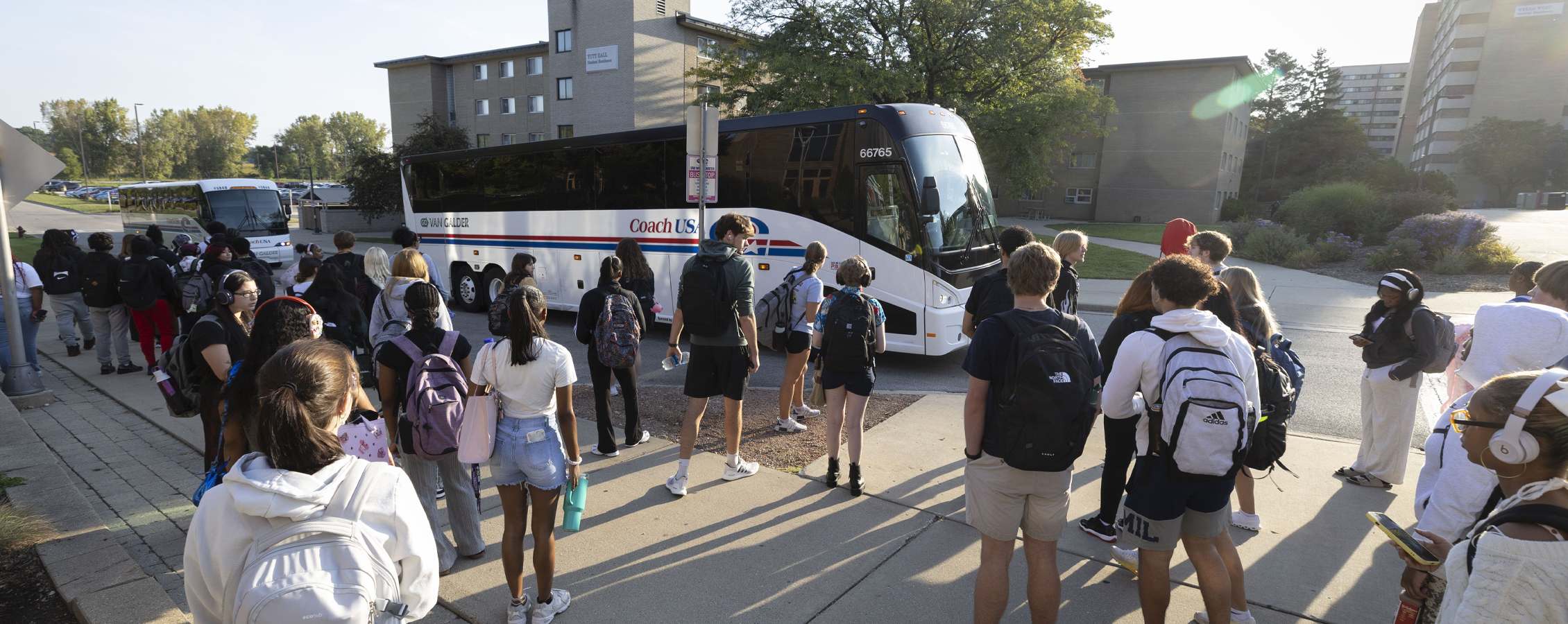 Two Coach buses pull up by the residence halls as students wait on the sidewalk.