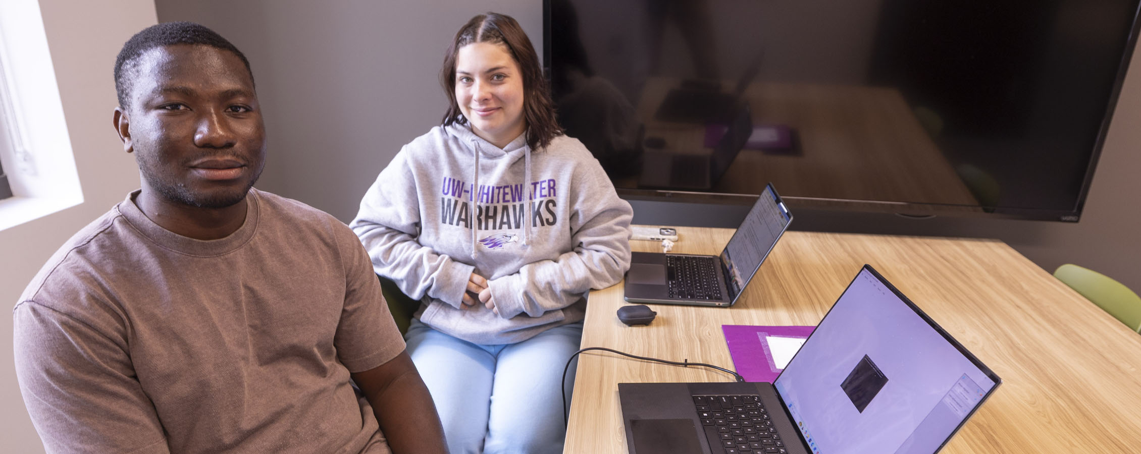 Two students sit at a table with laptops.