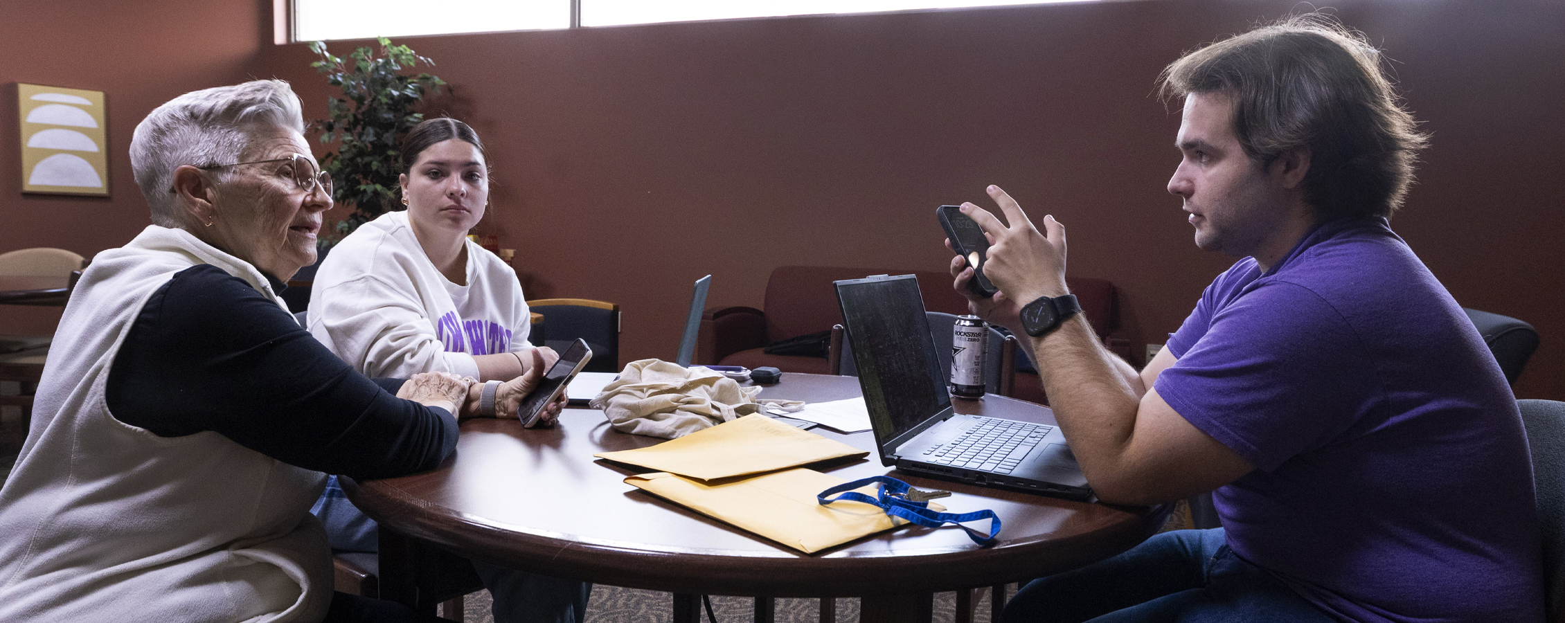 Two students with laptops talk to a senior citizen holding a smartphone.