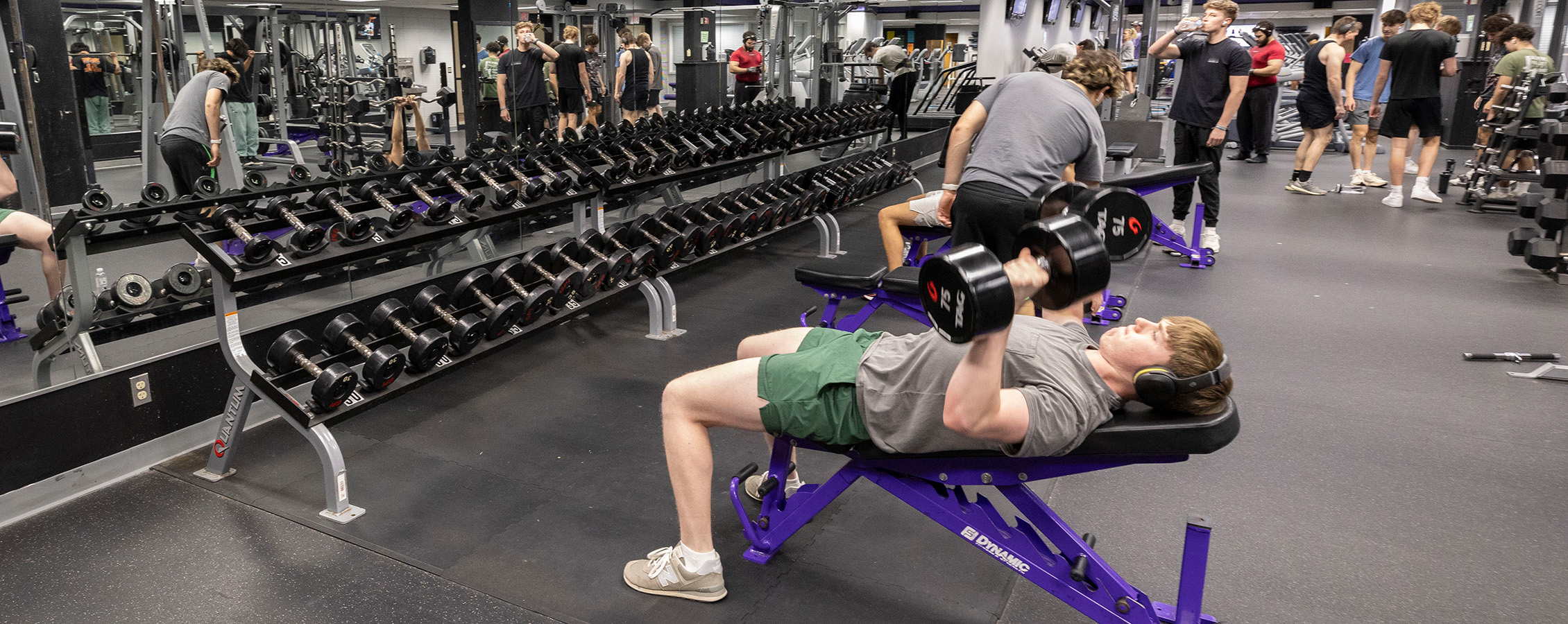 A student in the weight room does bench presses with other students working out in the background.