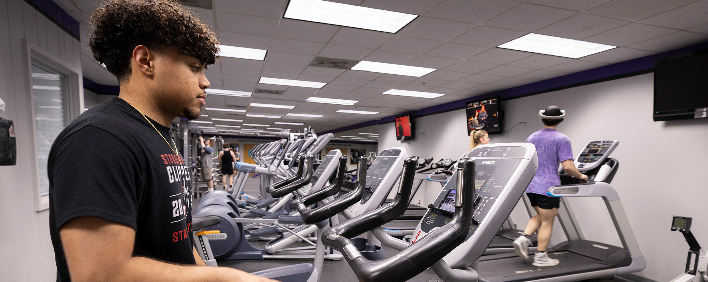 A student exercises in the University Fitness facility with another on a treadmill in the background.