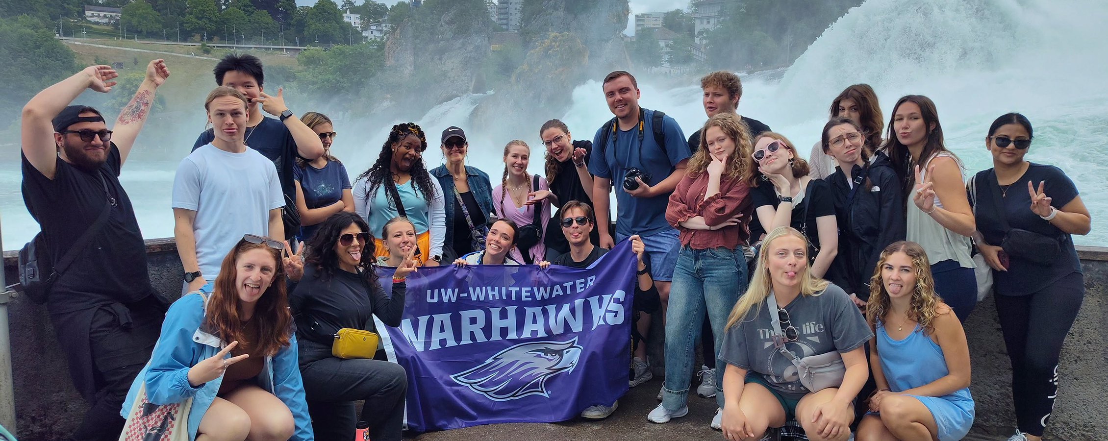 Communication students from UW-Whitewater group together for a photo while holding a UW-Whitewater Warhawks flag in front of a large water fountain.