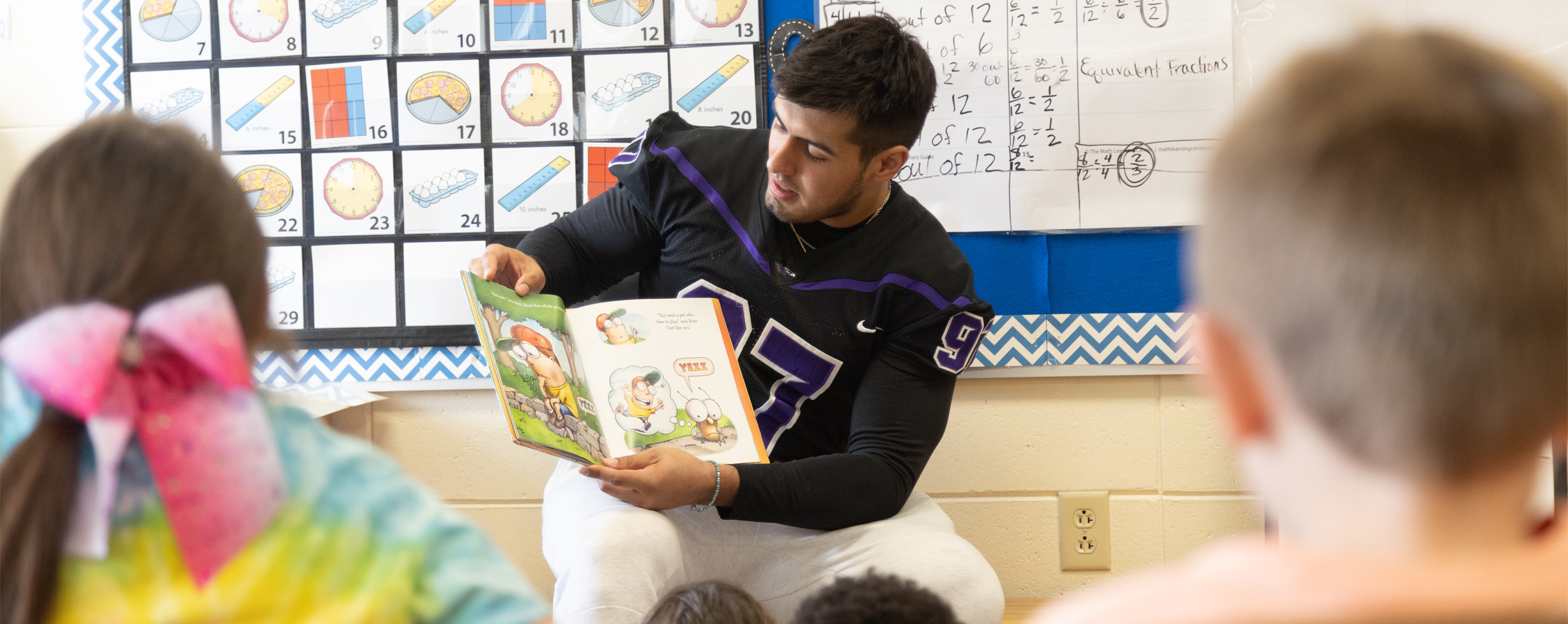 A UW-Whitewater student, wearing a Warhawk football jersery, reads a book to a classroom of children.