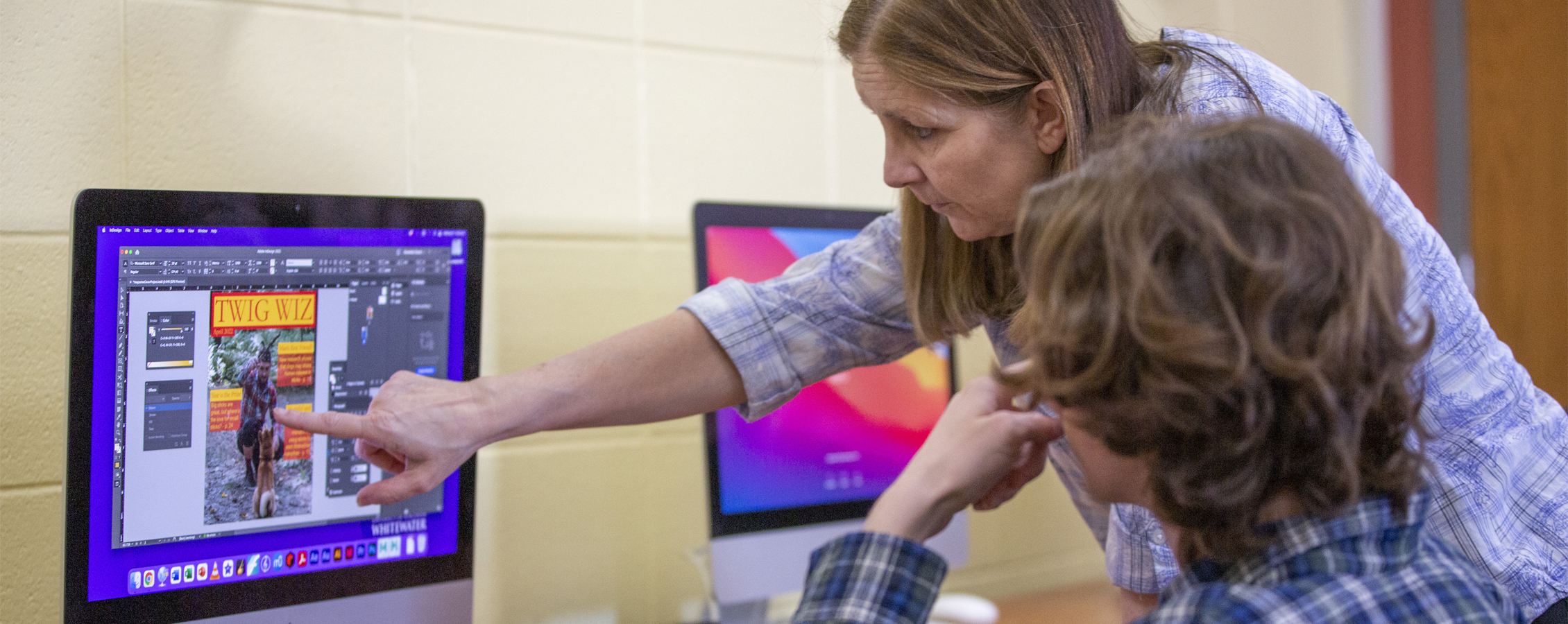 A faculty member points at a Mac computer as a student sits in a chair in front of the computer.