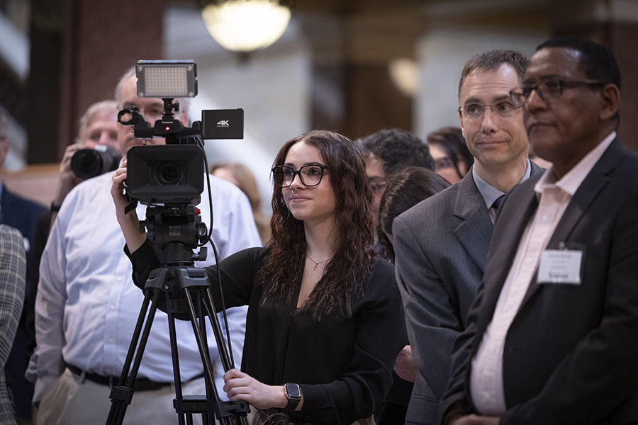 A student wearing a black blazer stands behind a camera in a crowd of people.