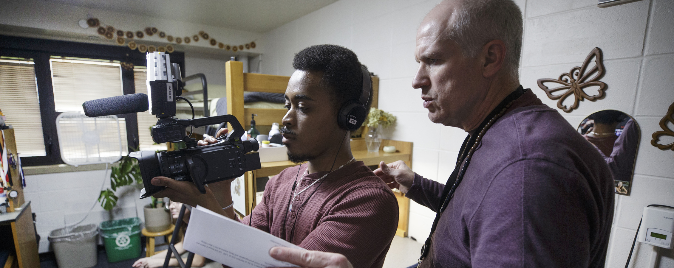 A student holds a video camera as a faculty member assists.
