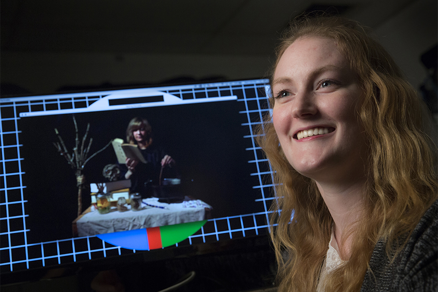 A student sits next to her work displayed on a computer.