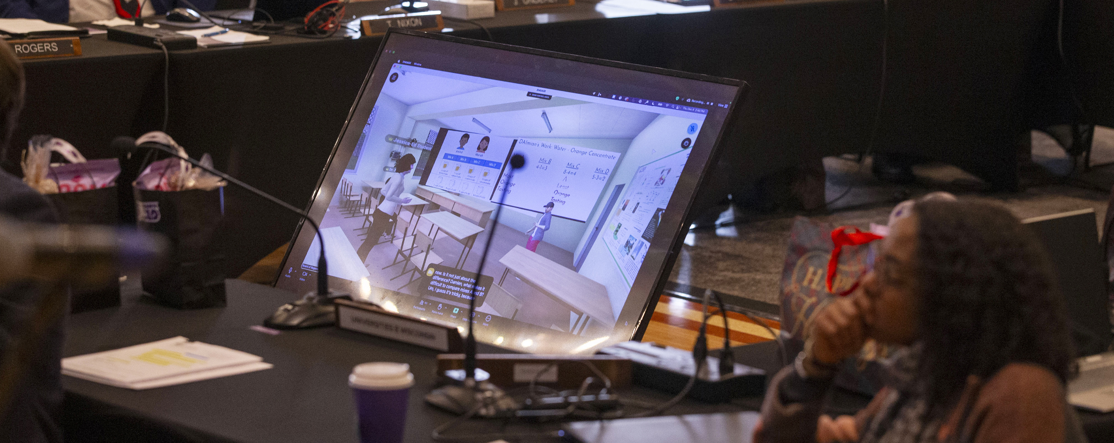 A person sits next to a screen monitor showing a classroom created by artificial intelligence.