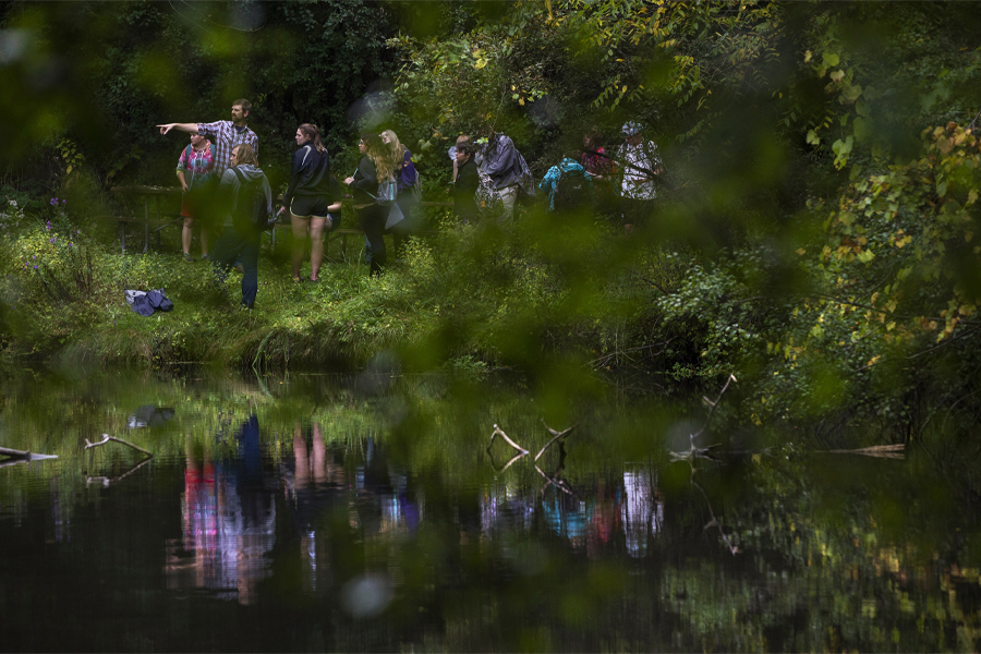 Students and a faculty member walk through a forest with a small body of water.