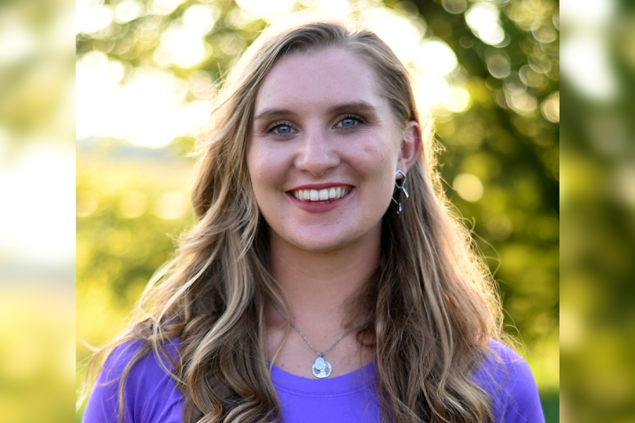 A person with long hair is wearing a blue shirt and standing outdoors with trees in the background.