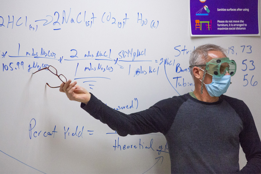 A faculty member points at the whiteboard while wearing safety goggles and a mask.