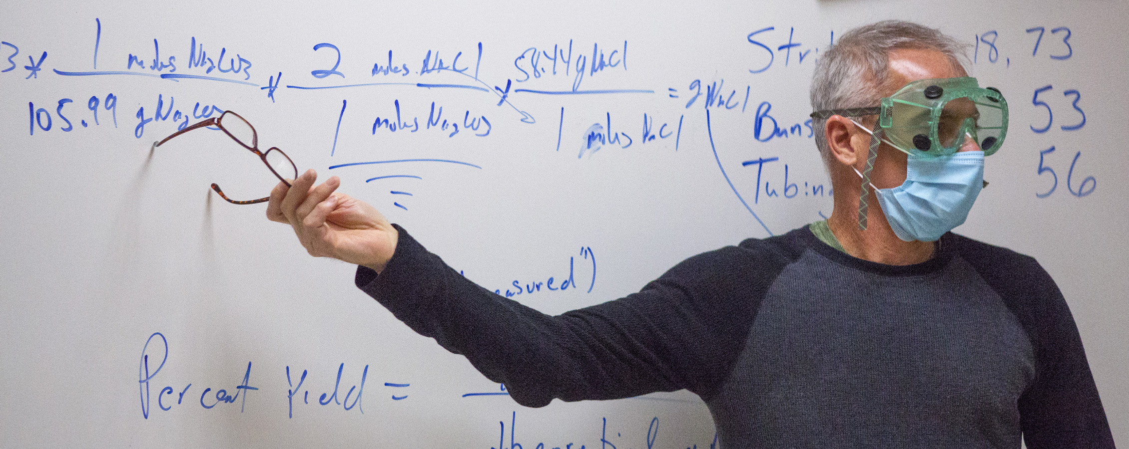 A faculty member, wearing safety googles and a face mask, points at a formula written on a whiteboard.