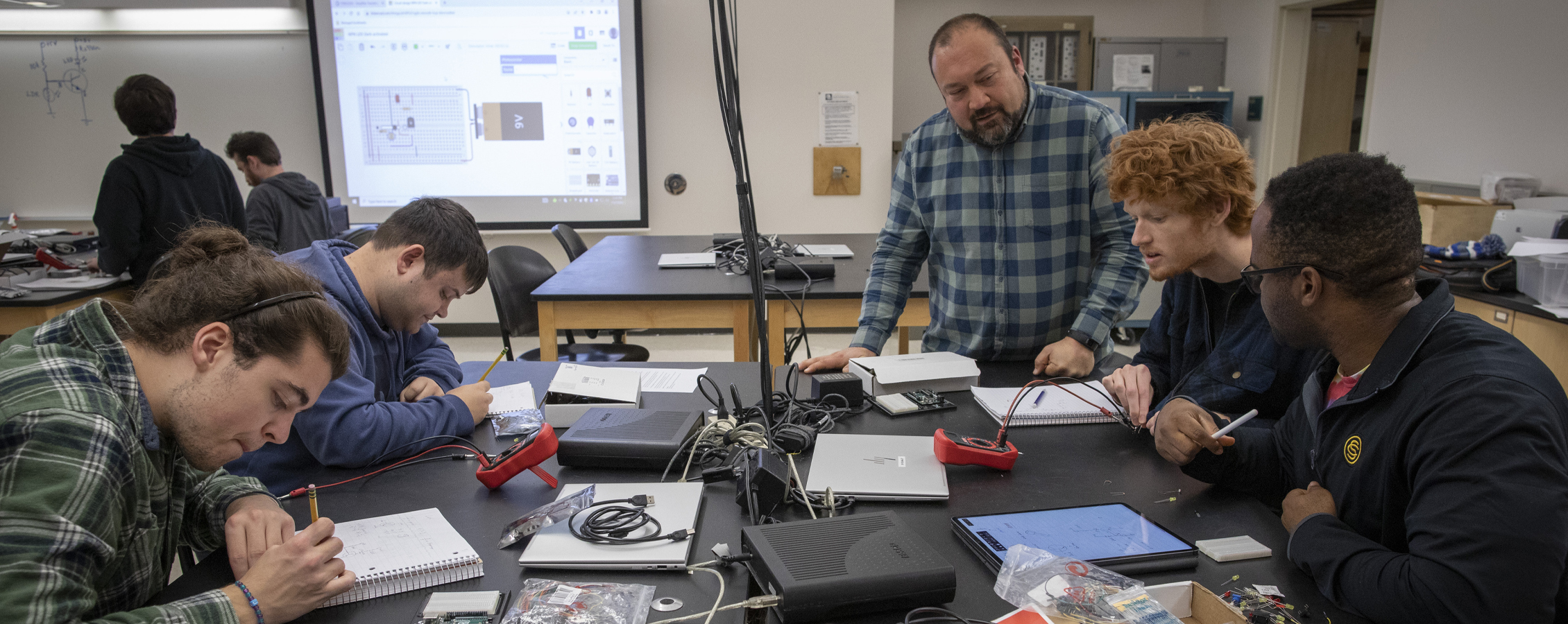 Students and a faculty member gather around a table as as they build circuitry. 