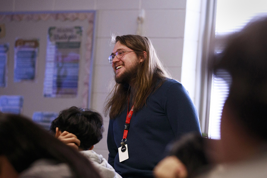 A person wearing a lanyard and a dark colored shirt smiles in a classroom.