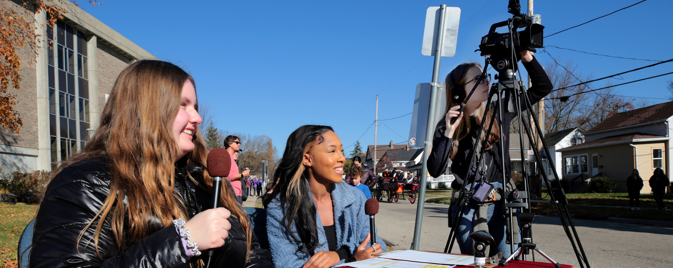 Two students hold microphones and one student works a video camera during a Homecoming parade.