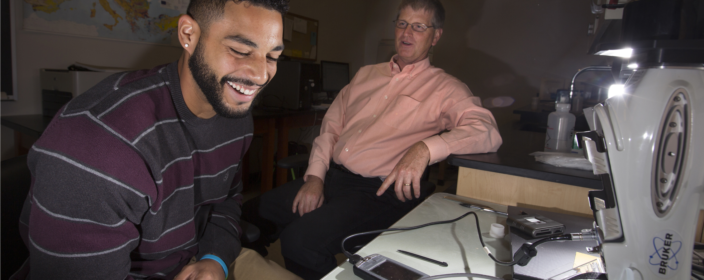 A student is indoors with his professor, using technology to test soil samples.