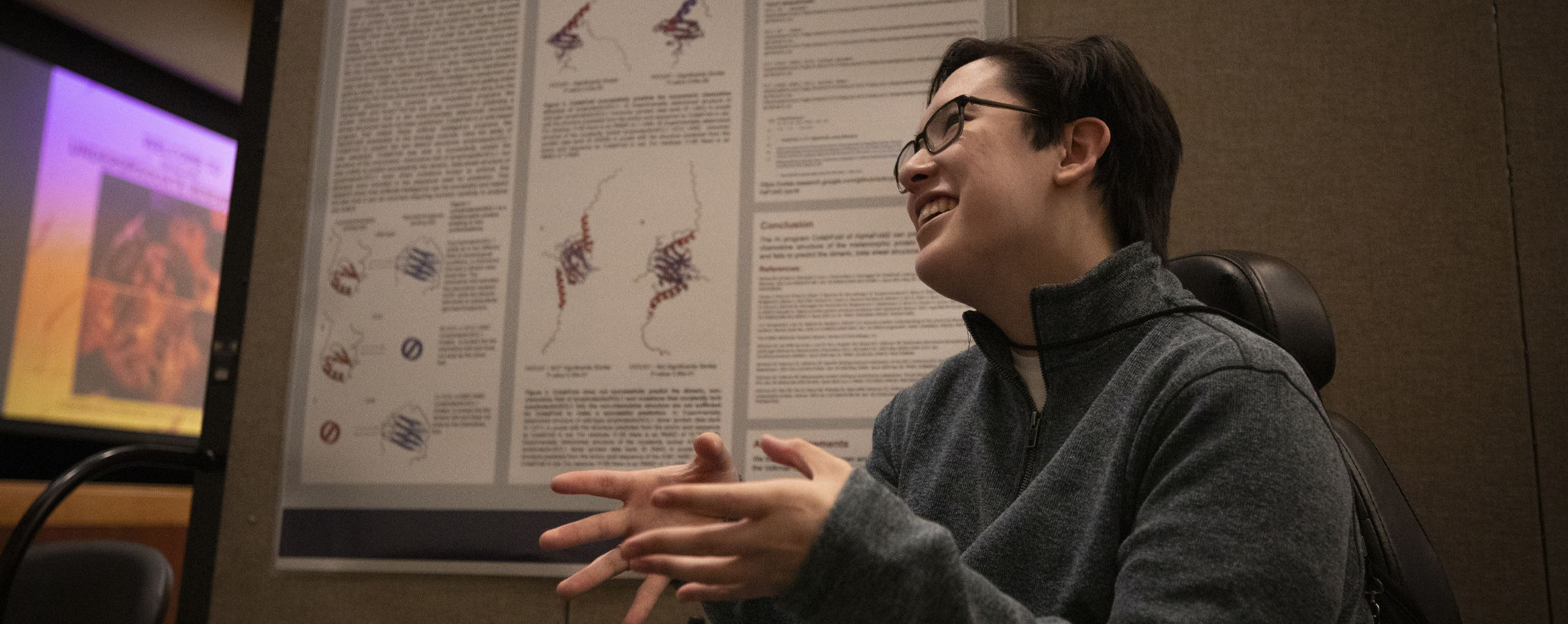 A student smiles and speaks with their hands in front of a research poster.