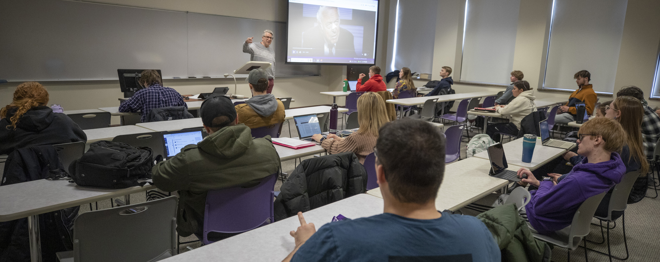 A faculty member points to a whiteboard at the front of class.