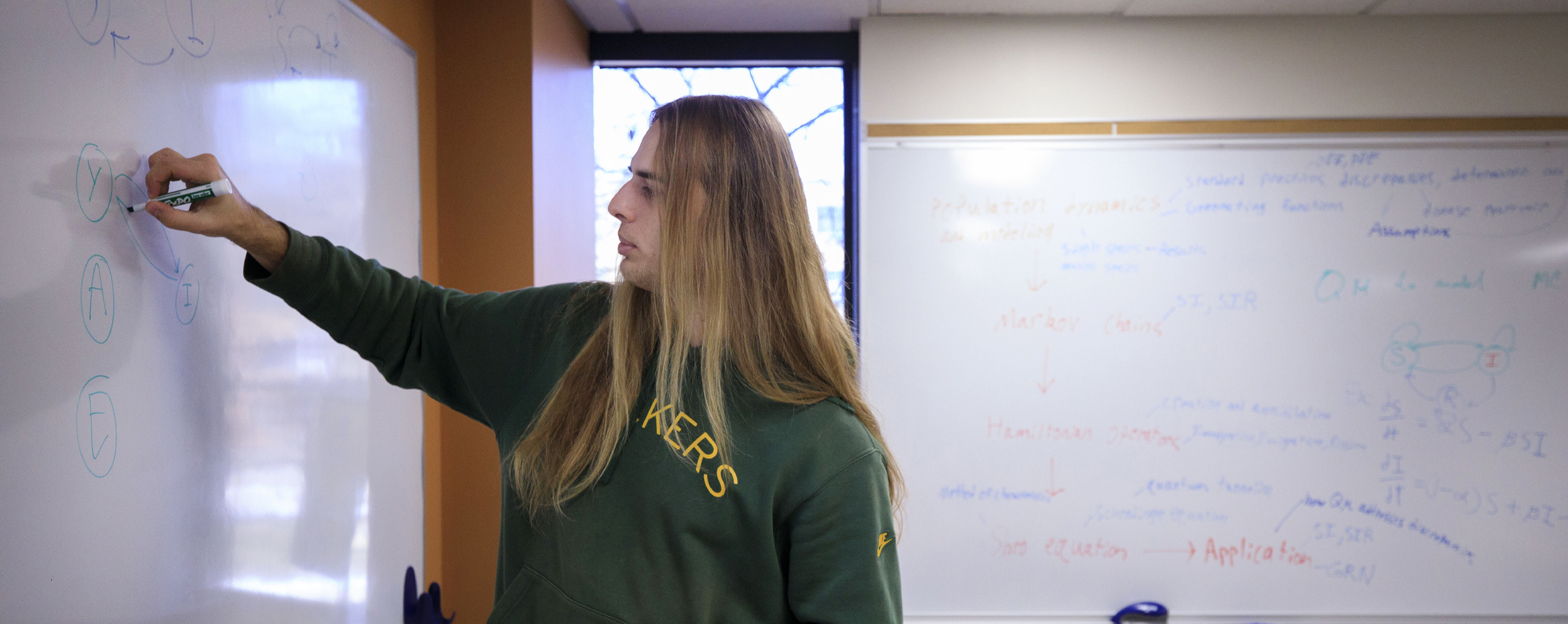 A student wearing a Green Bay Packers shirt writes on a whiteboard.