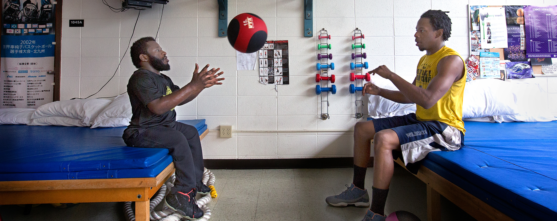 Two people toss a medicine ball while sitting down in a training room.