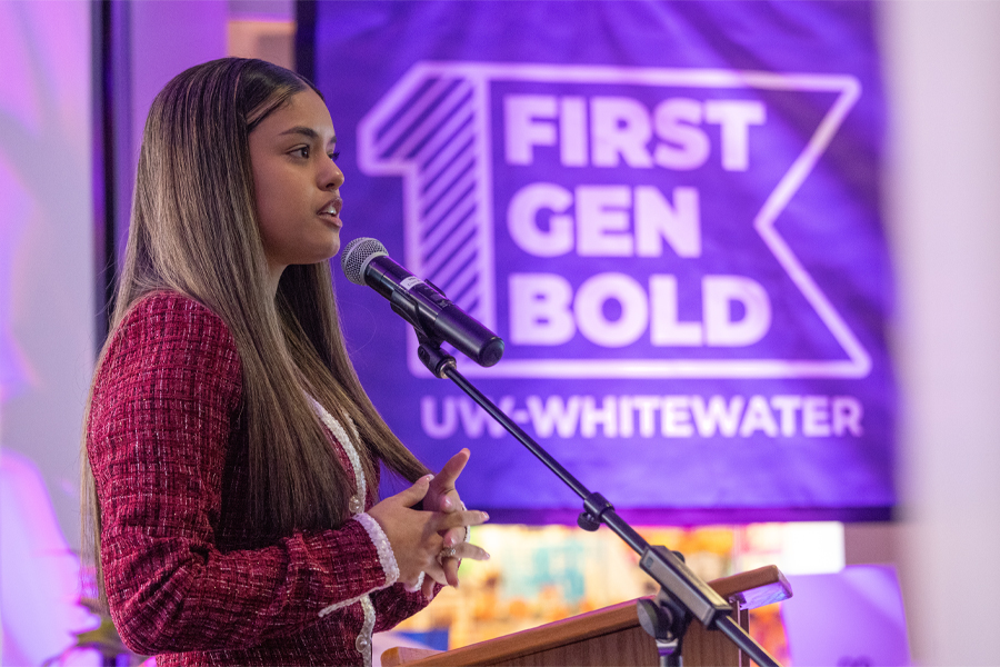 A student speaks at a podium with a purple flag in the background that says First Gen Bold.