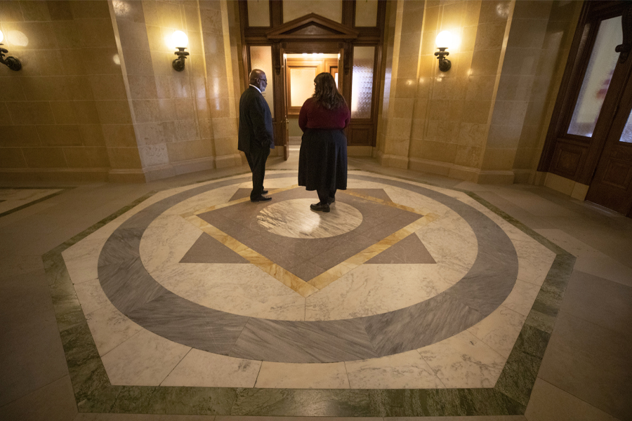 Two people talk in a room with dark woodwork and American flags.