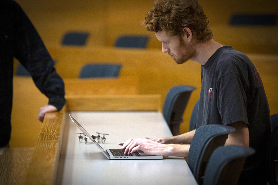 A student works on a laptop with a small drone sitting next to them on a table.