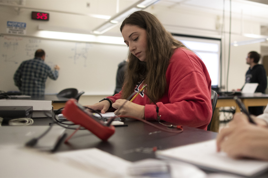 A student in a red shirt sits at a work table and takes notes on a notepad.