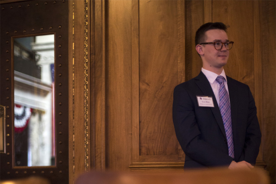 A student wears a suit and tie and stands against a wood paneled wall.