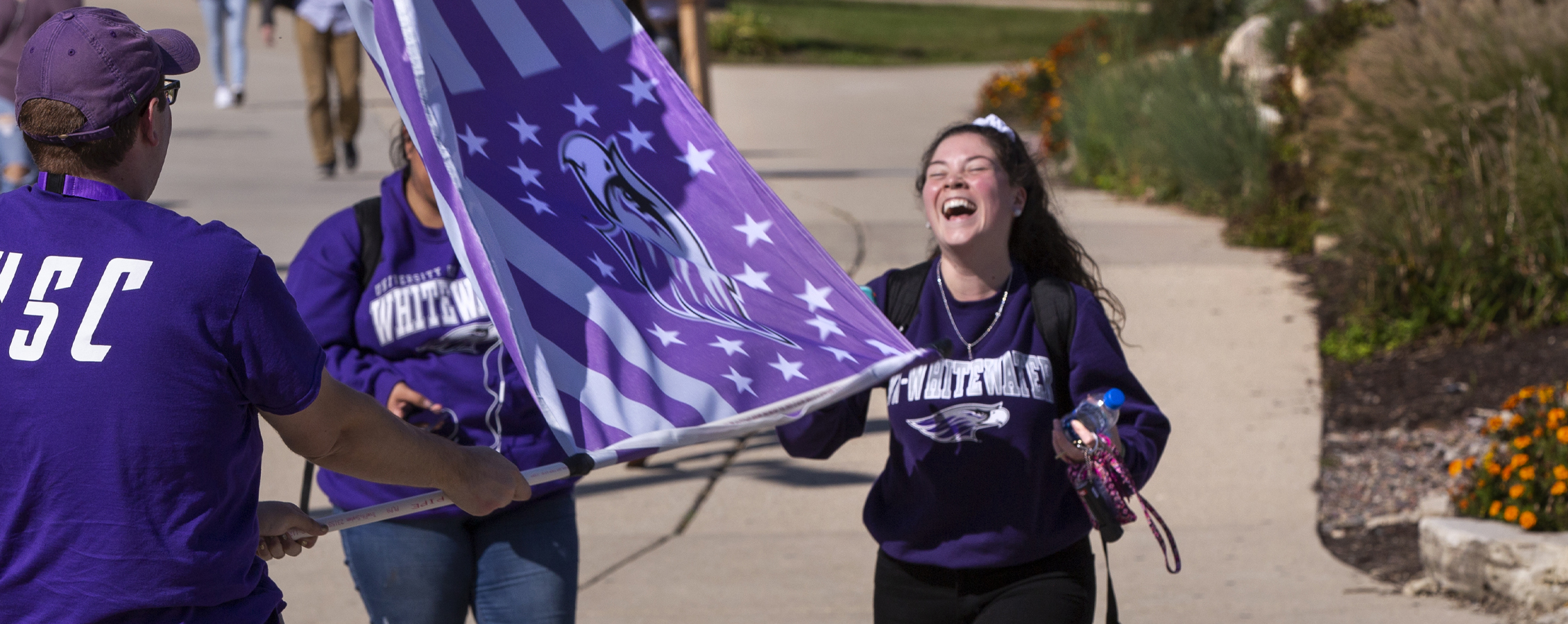 Three students laugh together, waving a purple Warhawk flag.