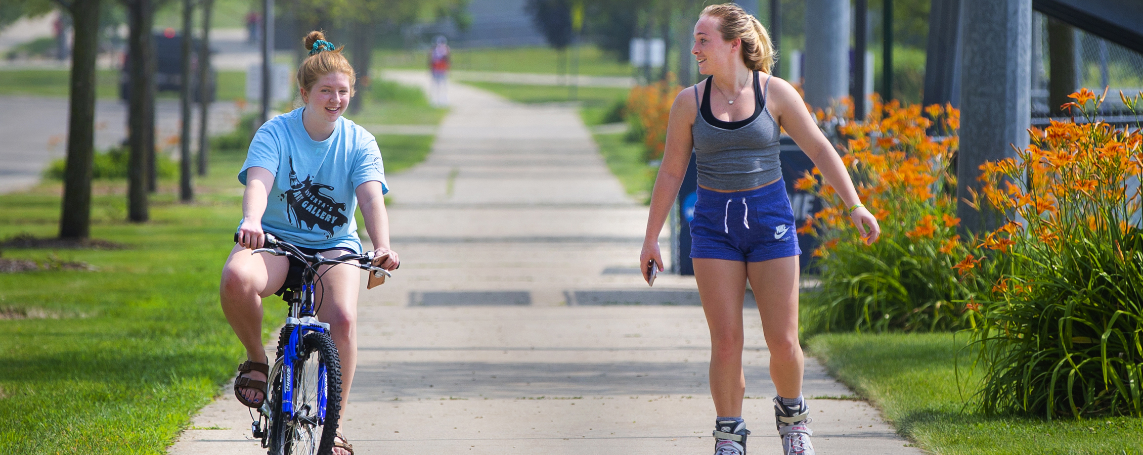 A student rides a bike next to another student rollerblading down a sidewalk.