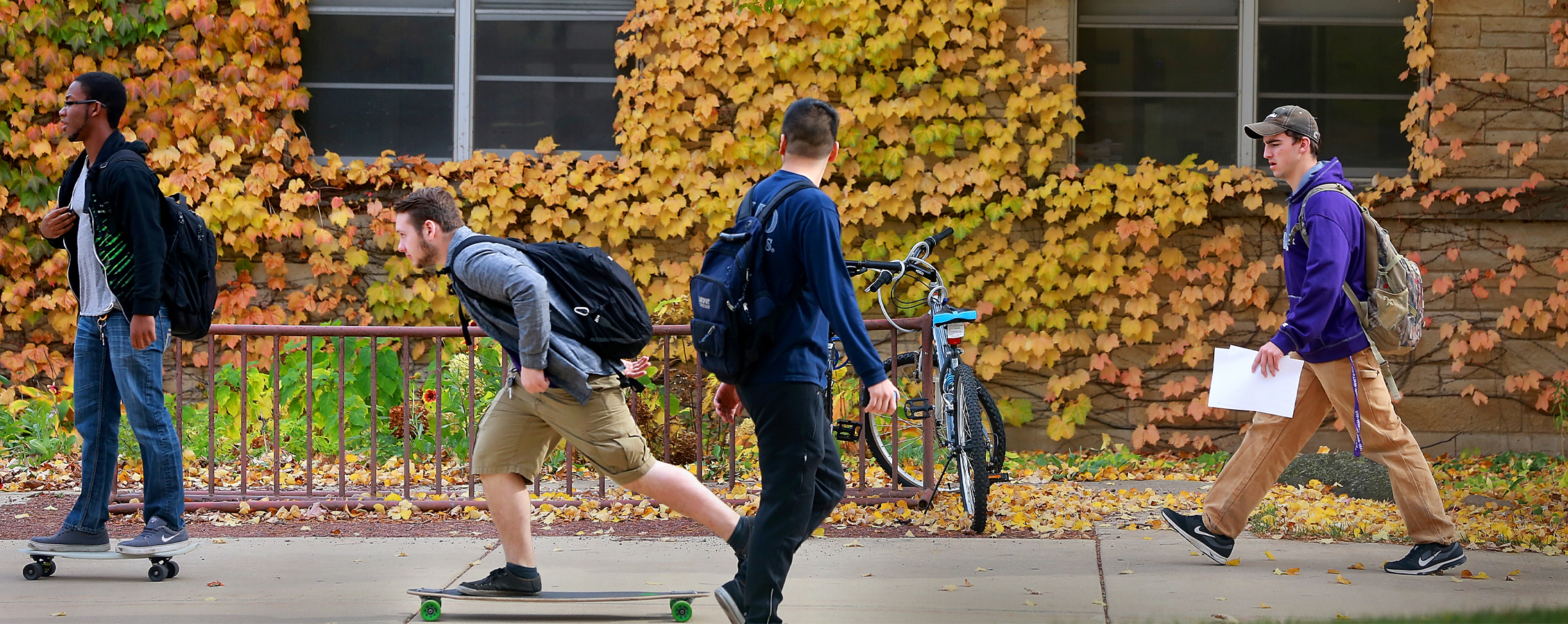 Students skateboard down a sidewalk with yellow foliage and fallen leaves in the background.
