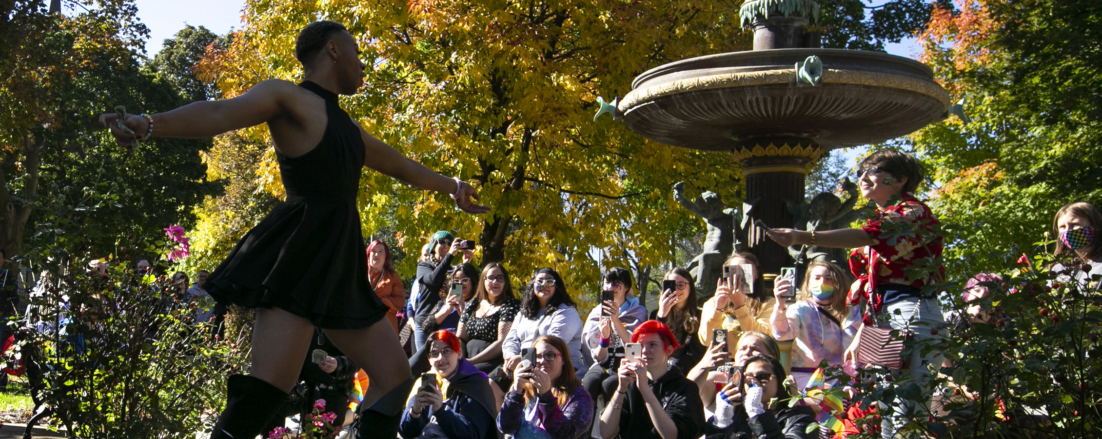 A person dances outdoors in front of a crowd by a water fountain.
