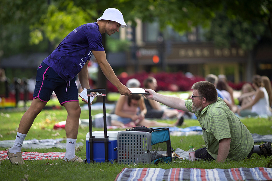 A student wearing a purple shirt hands a survey to a person having a picnic on a colorful blanket.