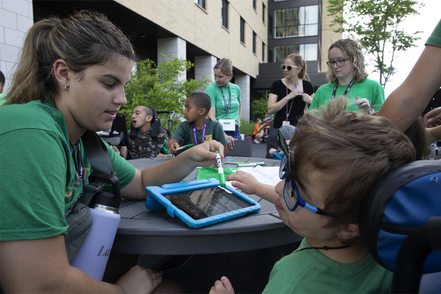 A student sits at a picnic table and interacts with a child who uses a device to communicate.