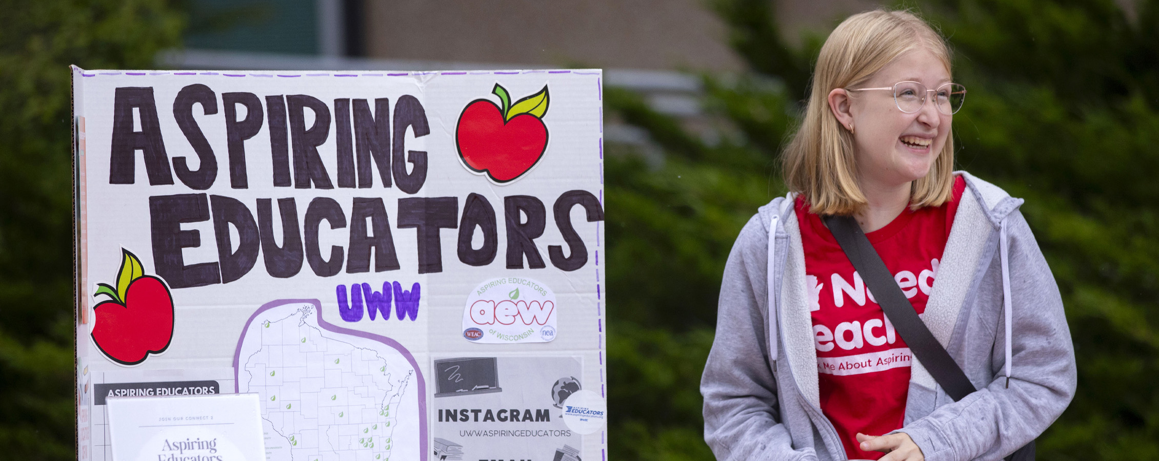 A smiling student stands at a table outdoors with a sign that says Aspiring Educators.