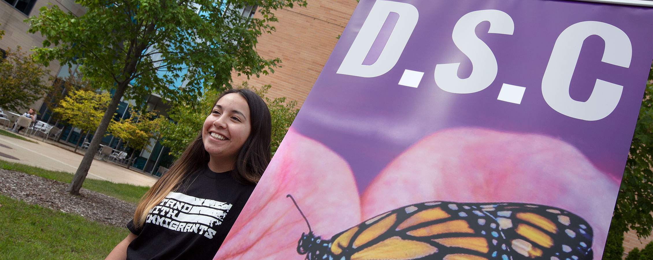 A student smiles and stands outdoors next to a sign that says D.S.C. with a monarch butterfly on it.