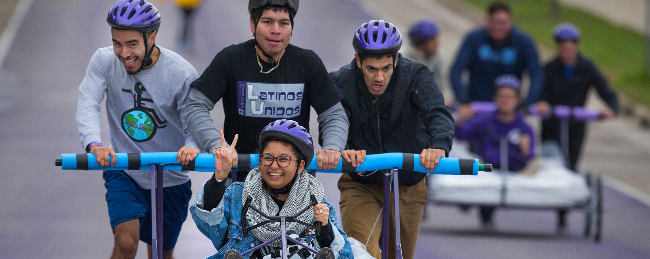A student group participates in bed races as three people push the bed and another rides with a smile on her face and giving the peace sign with two fingers.
